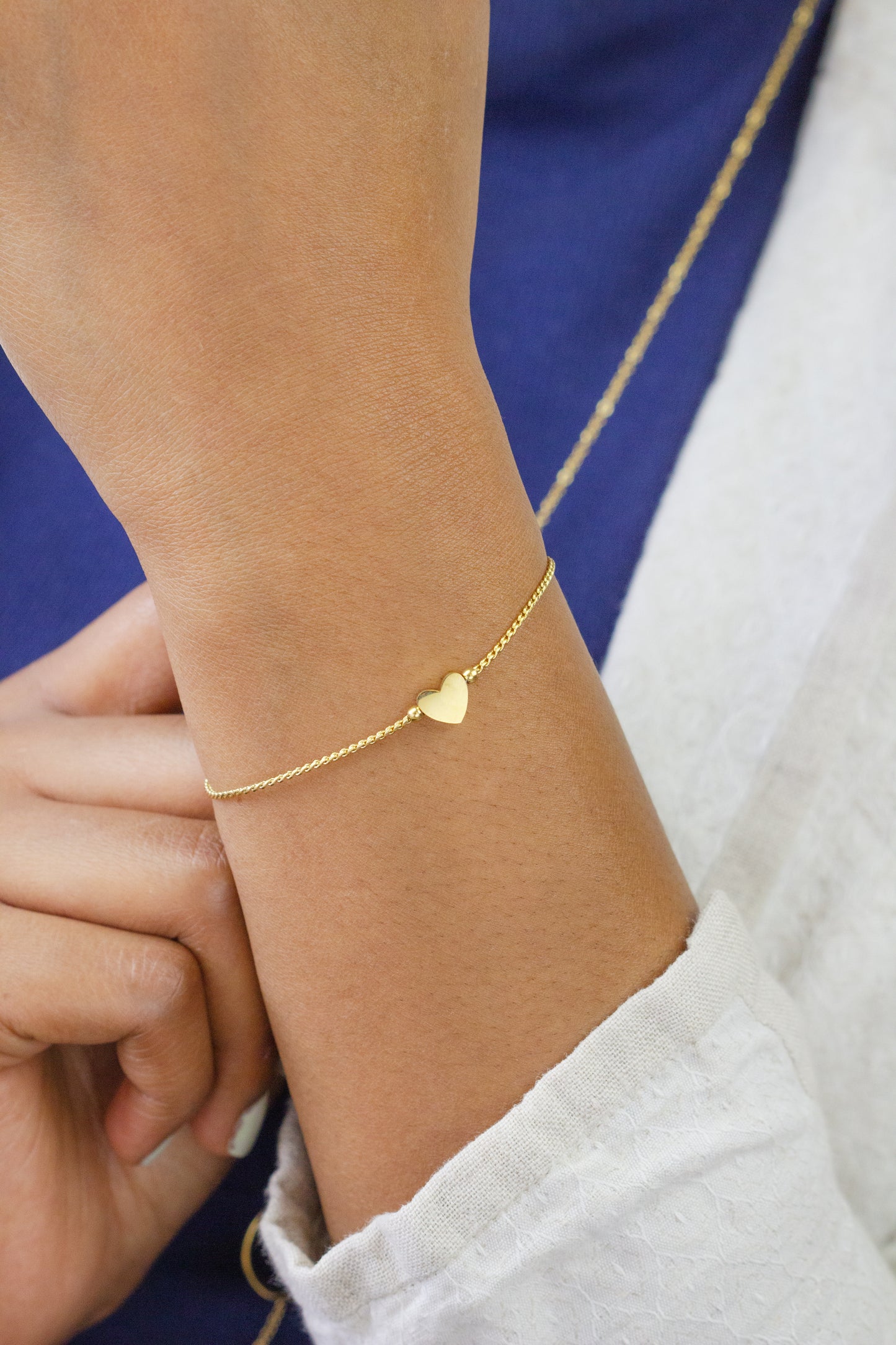 A close-up of a model's wrist adorned with a delicate gold bracelet featuring a small heart charm from Dahlia Jewels. The model is wearing a blue top and white outerwear. Dahlia Jewels offers free delivery in Nairobi.