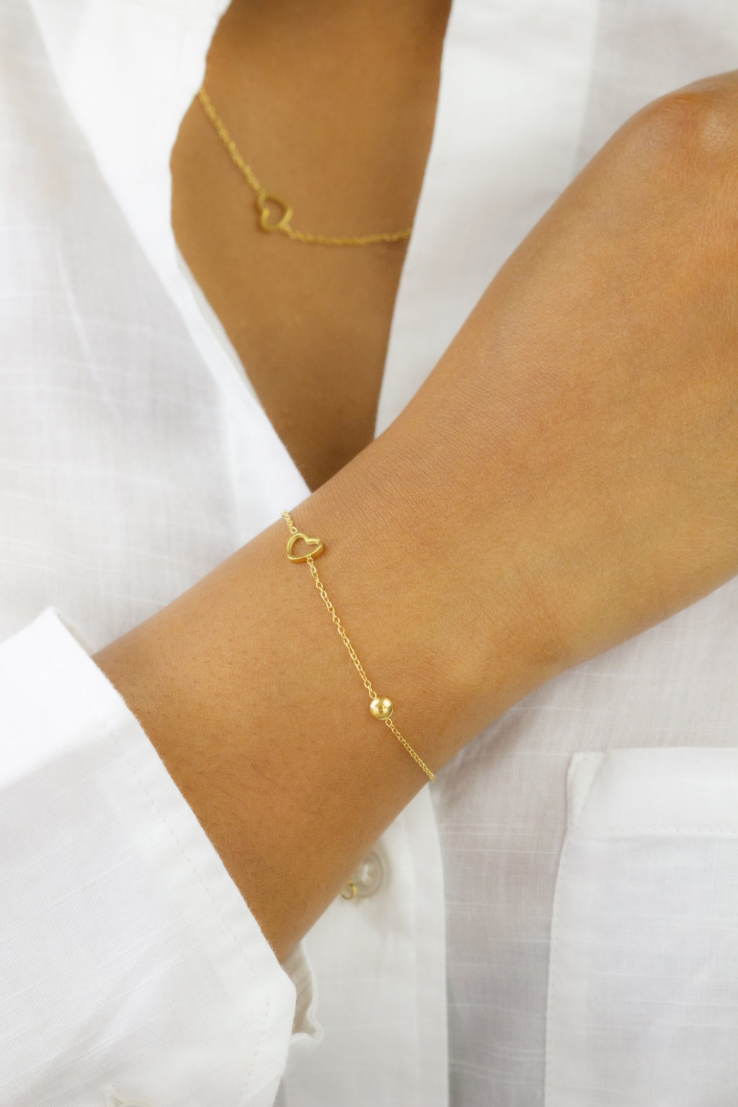 A close-up of a model's arm adorned with a delicate gold bracelet featuring a heart charm from Dahlia Jewels. The model wears a white shirt, and part of a matching gold necklace is visible. Dahlia Jewels offers free delivery in Nairobi.