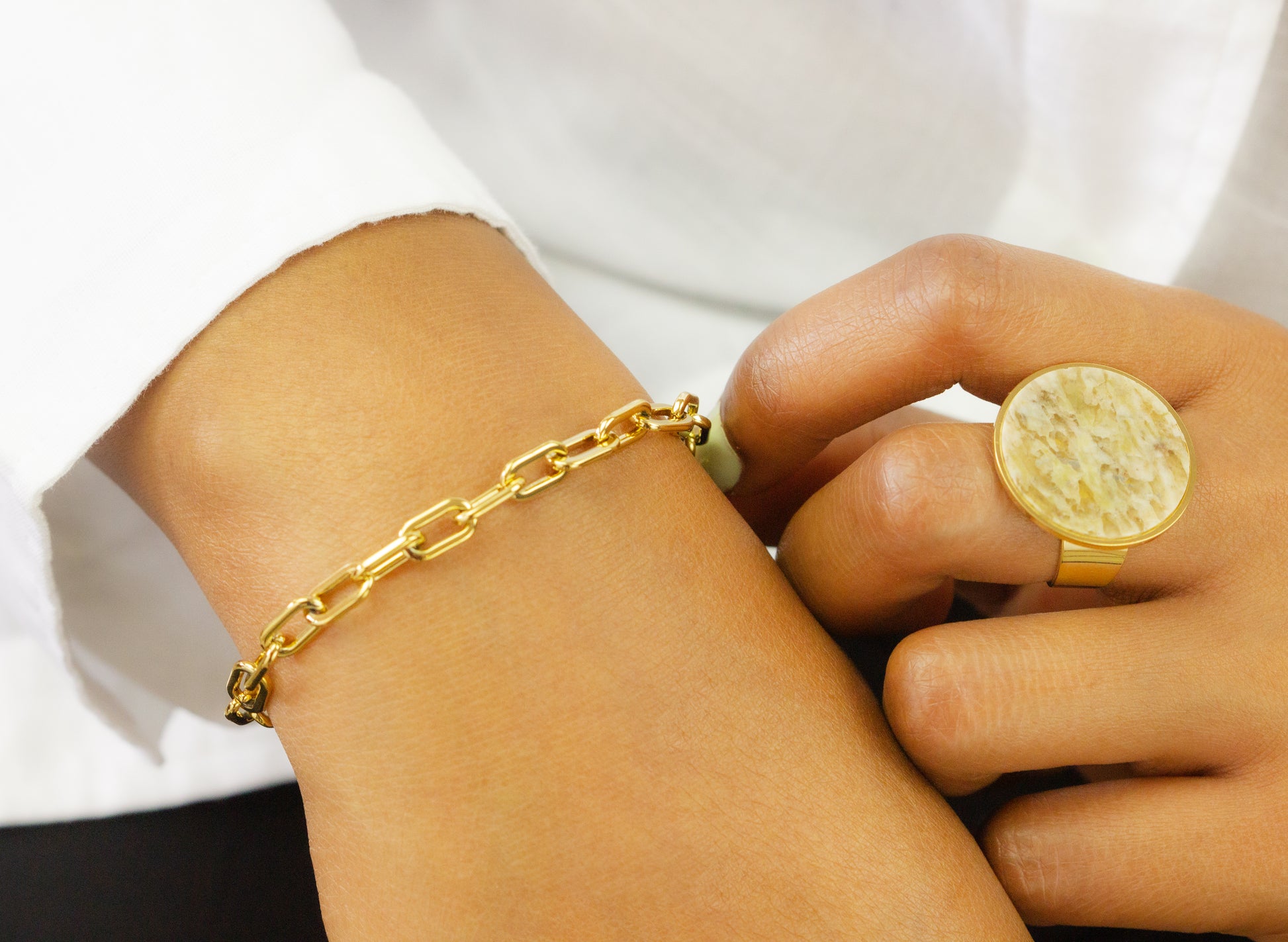 A close-up of a model's wrist adorned with a chunky gold chain bracelet and a large round stone ring from Dahlia Jewels. The model is wearing a white sleeve. Dahlia Jewels offers free delivery in Nairobi.