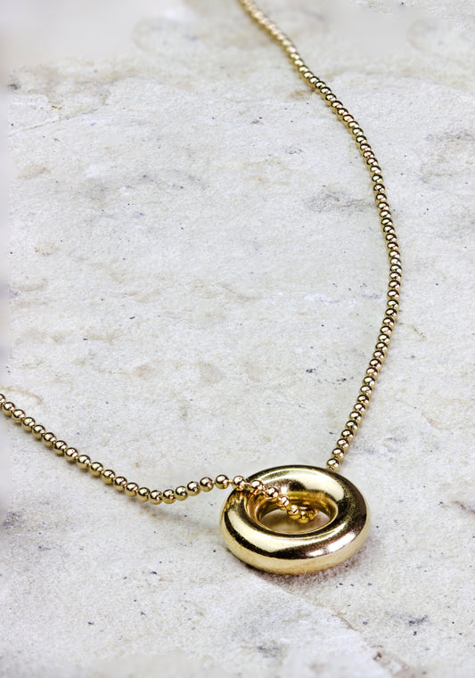 Striking gold-plated necklace from Dahlia Jewels, showcasing a bold circular pendant with a spiral design that captures a sense of movement and sophistication. This pendant is suspended on a fine chain made of small, round beads that complement its glossy finish. Ideal for adding a modern twist to any outfit, this necklace is versatile enough for both everyday wear and special occasions. Secures with a standard clasp. Available for free delivery in Nairobi. Keywords: jewellery, jewelry.