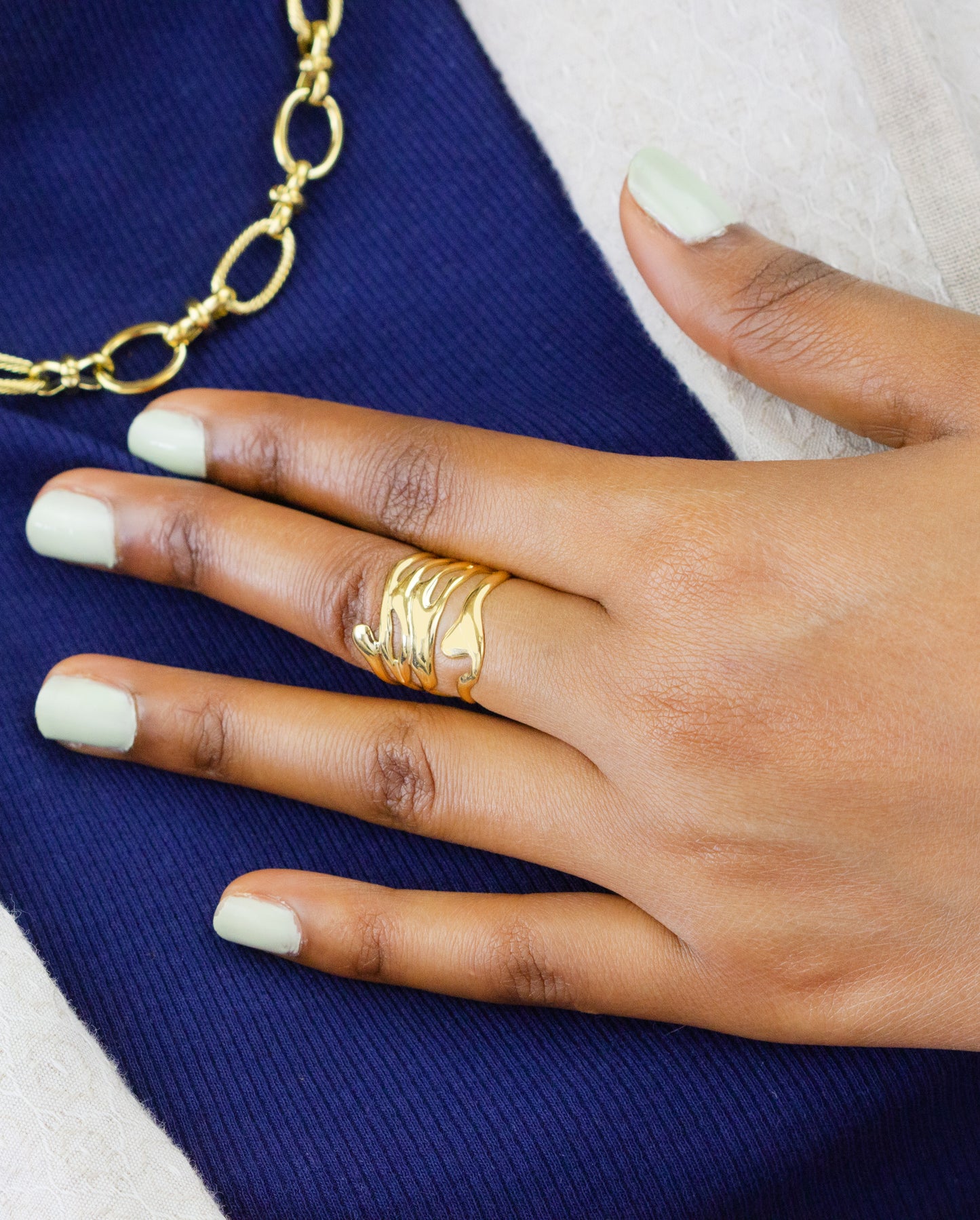 A close-up of a model's hand featuring a unique gold ring with a wave design from Dahlia Jewels. The model's fingers are adorned with a light green nail polish, complementing the gold jewelry. The image also shows a part of a gold necklace. Dahlia Jewels offers free delivery in Nairobi.