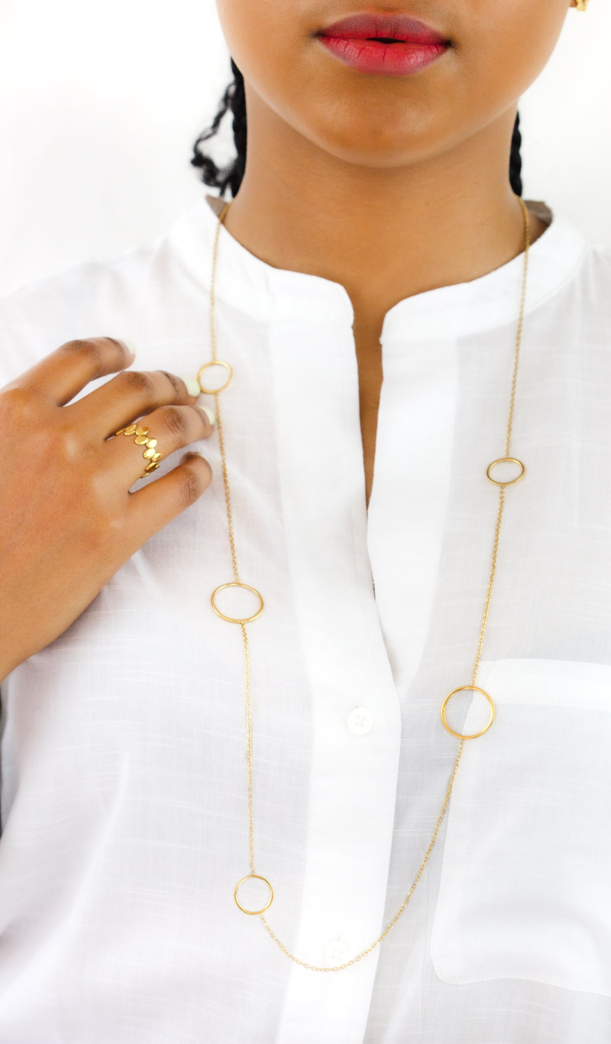 A model wearing a long gold necklace with circular rings and a matching gold ring from Dahlia Jewels. The minimalist and elegant design is complemented by the model's white blouse. Dahlia Jewels offers free delivery in Nairobi.