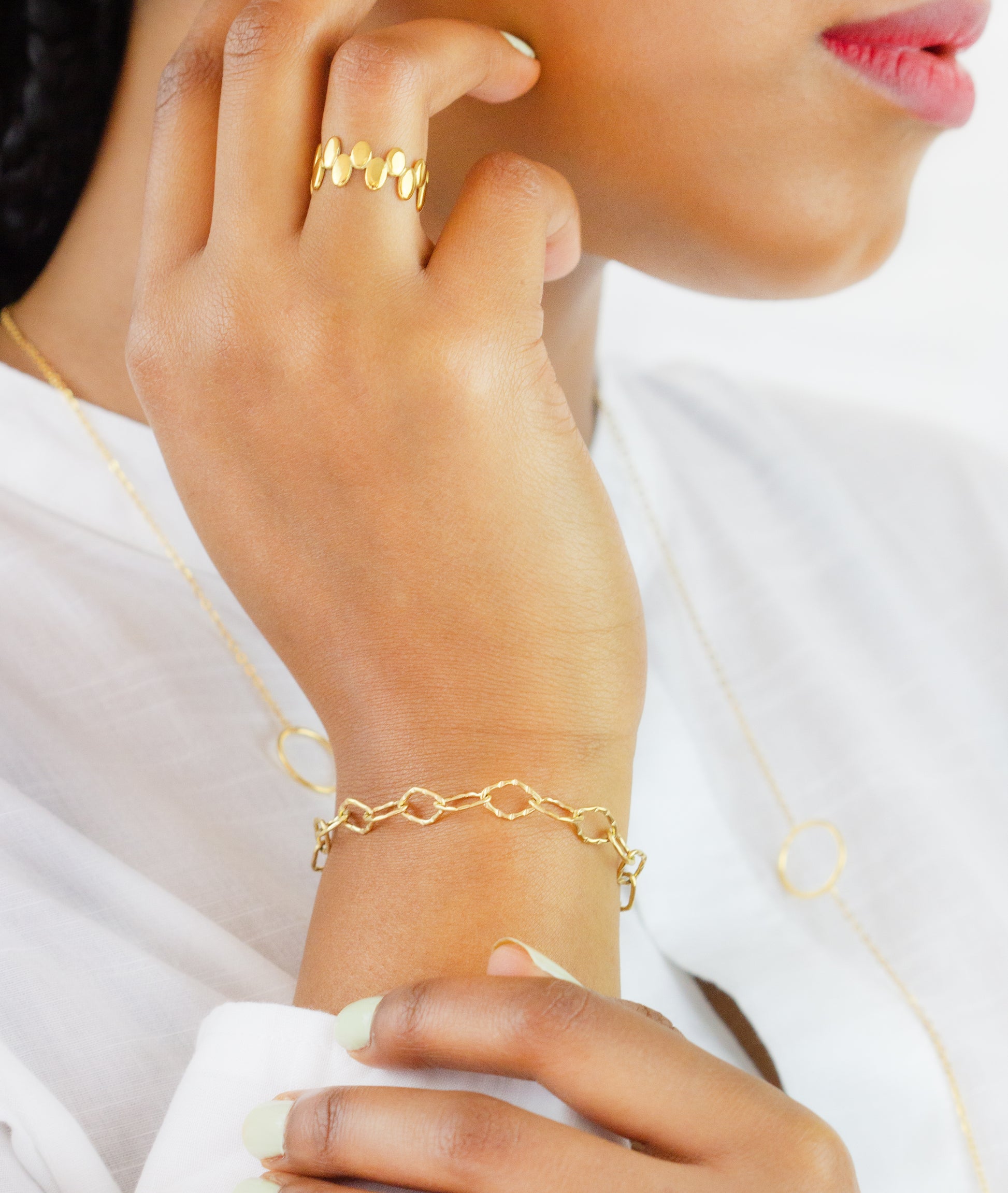 A close-up of a model's hand showcasing a gold bracelet with an elegant chain link design and a gold ring with circular details from Dahlia Jewels. The model is wearing a white blouse and a matching gold necklace is partially visible. Dahlia Jewels offers free delivery in Nairobi.