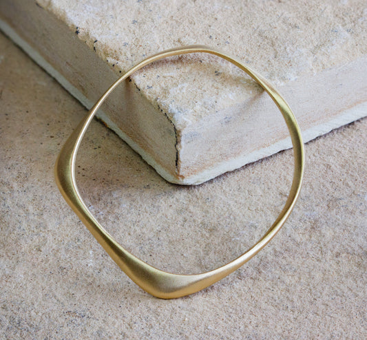 Minimalist gold bangle bracelet with a modern, asymmetric design, displayed on a textured stone surface. The bangle features a smooth, matte finish, highlighting its elegant and contemporary aesthetic. Available with free delivery in Nairobi. #gorgeouseveryday