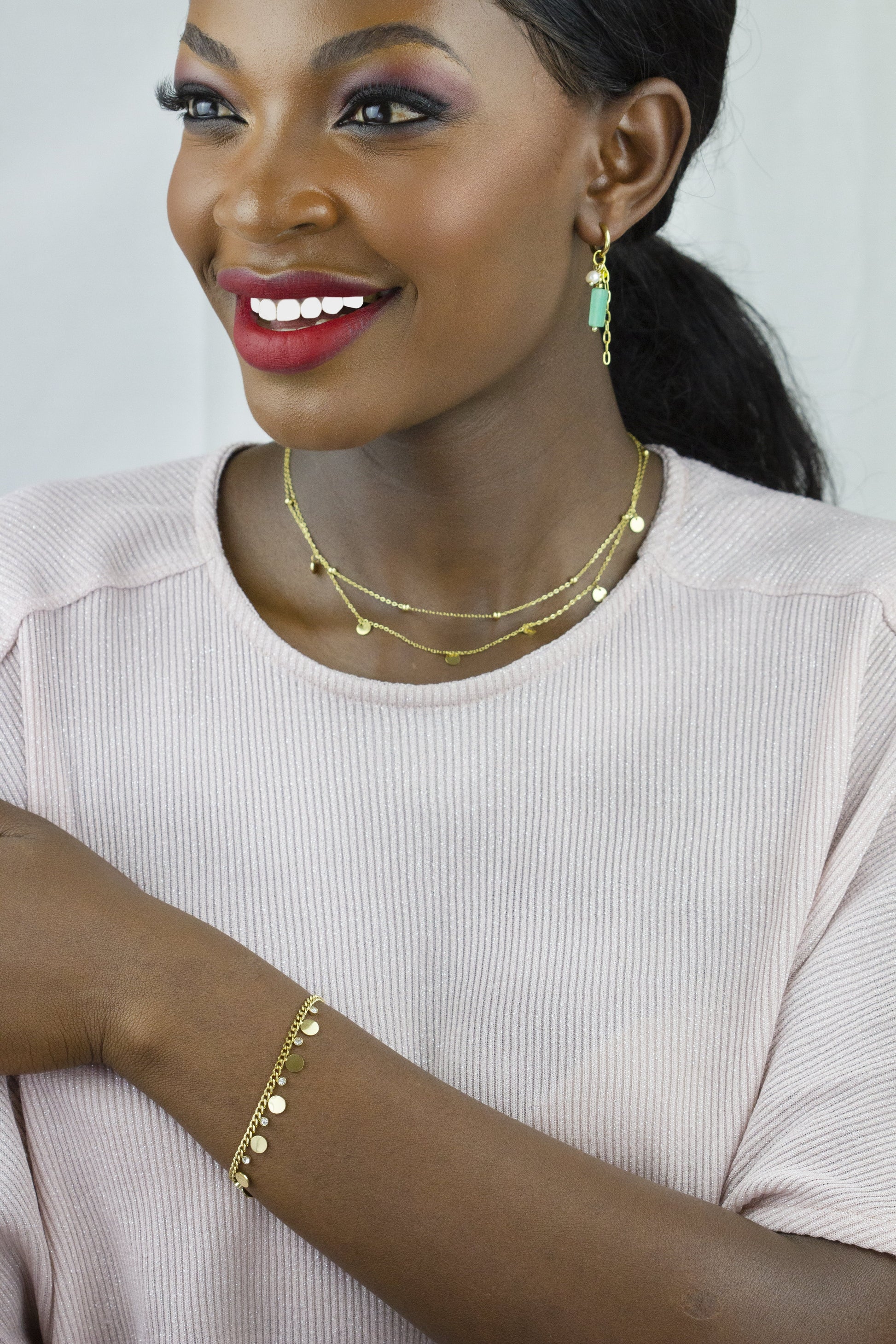 The model showcases a gold-tone bracelet from Dahlia Jewels on her arm, complemented by a layered gold necklace with small discs, adding a sophisticated touch to her look. The jewelry pairs perfectly with her attire. Dahlia Jewels offers free delivery in Nairobi.