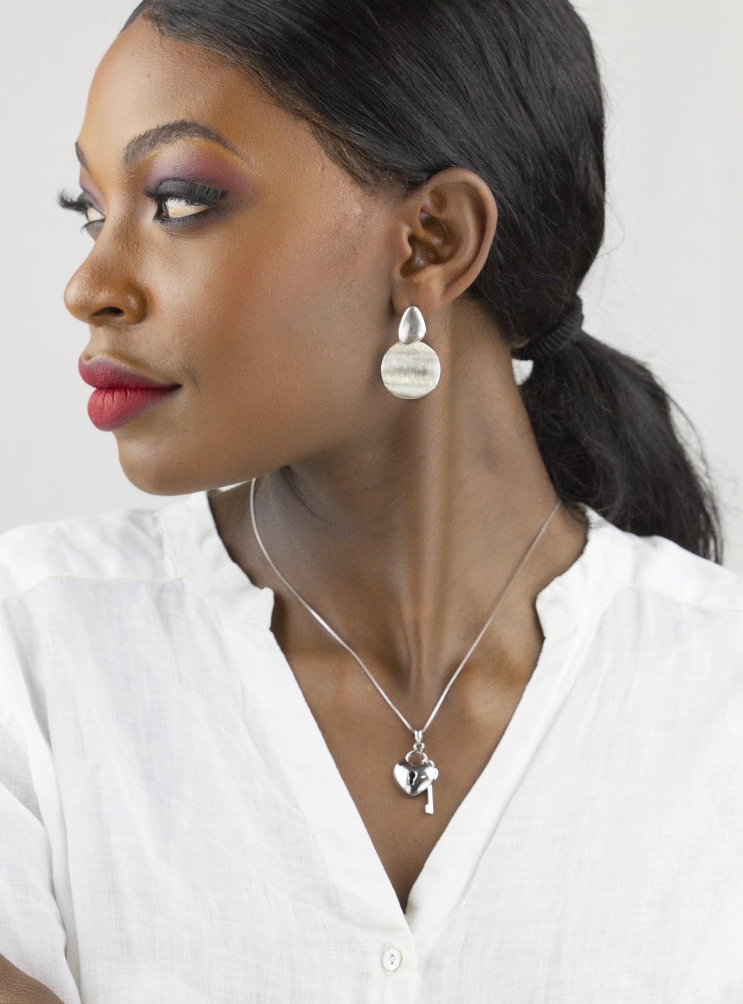 In a pristine white setting, Dahlia Jewels' model showcases silver disk earrings and a heart key pendant necklace, exuding sophistication and grace. Indulge in the luxury of jewellery with the convenience of free delivery in Nairobi.