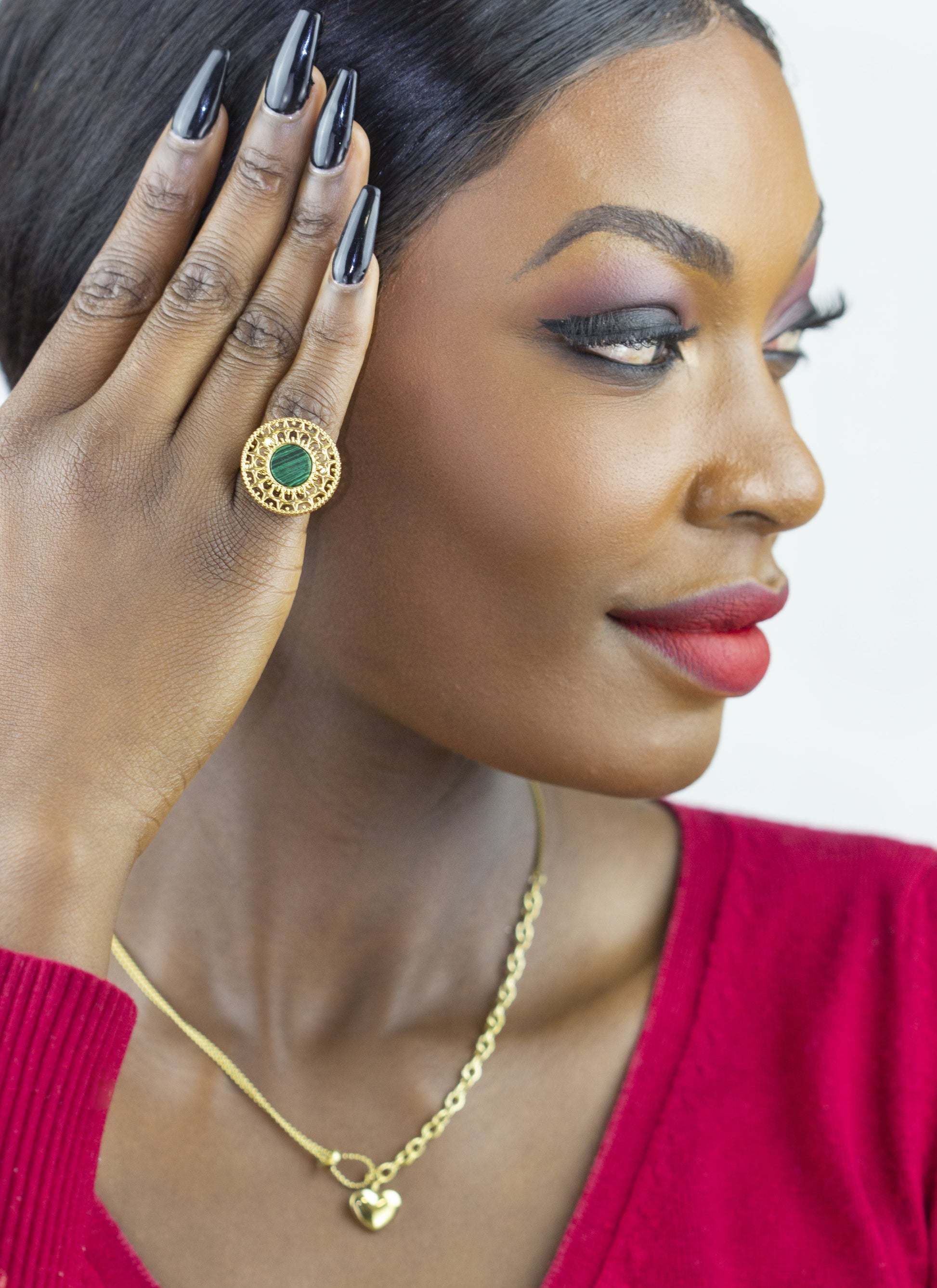 A model showcases Dahlia Jewels' exquisite craftsmanship, wearing a statement ring with a central green gem encircled by a golden patterned halo. Complementing the ring is a delicate necklace with a tiny heart pendant. Both items embody the brand's commitment to elegant jewellery/jewelry, available with free delivery in Nairobi.