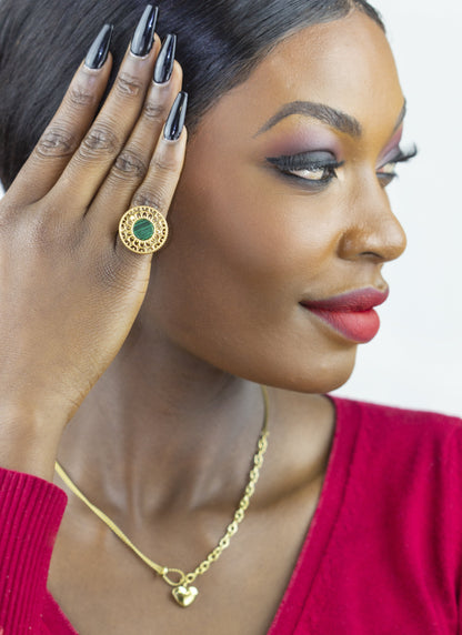 A model showcases Dahlia Jewels' exquisite craftsmanship, wearing a statement ring with a central green gem encircled by a golden patterned halo. Complementing the ring is a delicate necklace with a tiny heart pendant. Both items embody the brand's commitment to elegant jewellery/jewelry, available with free delivery in Nairobi.